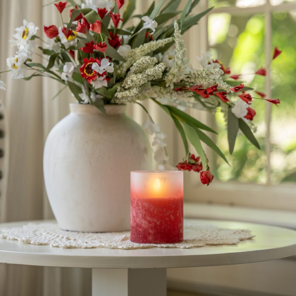 Red flameless candle beside a white vase of red and white winter stems on a lace doily